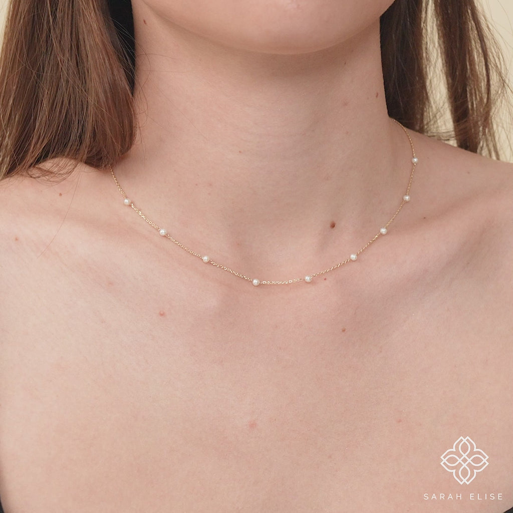 Model wearing a gold natural pearl station necklace, layered with other delicate necklaces.